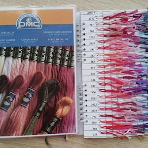 May include: A spiral-bound DMC embroidery floss color chart and guide. The left side displays a variety of floss skeins in shades of pink, red, and brown. The right side shows color swatches with corresponding DMC numbers. The chart includes text in English and French.