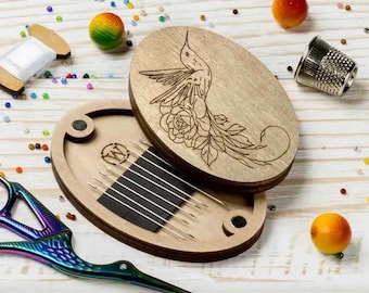 May include: A wooden needle minder with a hummingbird and floral design on the lid. The minder has a magnetic closure and holds a set of needles in the bottom compartment.