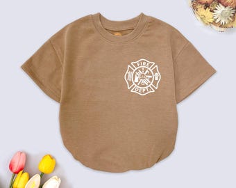 May include: A brown short-sleeved romper featuring a white "Fire Dept." emblem. The romper has a crew neck and a rounded hem. The image is taken from above, with flowers in the background. The romper is designed for babies or toddlers.