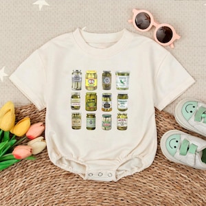 May include: Cream-colored baby bodysuit featuring a colorful graphic of various pickle jars. The jars are arranged in rows and columns, showcasing different brands and styles of pickles. The design is playful and food-themed.