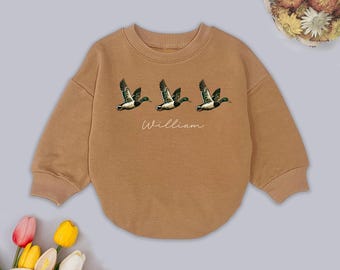 Może przedstawiać: Brązowa bluza z nadrukiem trzech lecących kaczek w kolorach zielonym, białym i czarnym. Imię "William" jest napisane kursywą pod kaczkami. Bluza ma okrągły dekolt i długie rękawy.