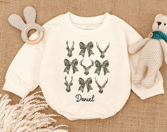 May include: Cream-colored baby onesie with long sleeves, featuring a pattern of deer antlers and bows in a camouflage design. The name "Daniel" is printed in black script at the bottom. A baby toy and stuffed bear are in the background.