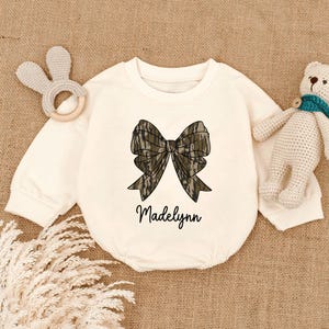 May include: Cream-colored baby onesie with a large bow graphic in shades of brown and green camouflage. The name "Madelynn" is written in a cursive font below the bow. A bunny-shaped rattle and a teddy bear are visible.