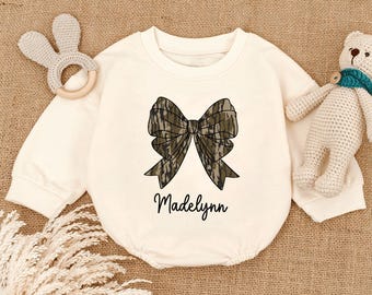 May include: Cream-colored baby onesie with a large bow graphic in shades of brown and green camouflage. The name "Madelynn" is written in a cursive font below the bow. A bunny-shaped rattle and a teddy bear are visible.