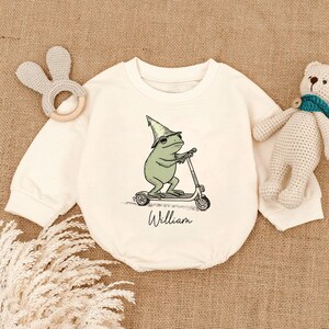 May include: Cream-colored baby onesie featuring a green frog riding a scooter and wearing a wizard hat. The name "William" is written in script below the frog. A bunny-shaped teether and a teddy bear are also in the image.