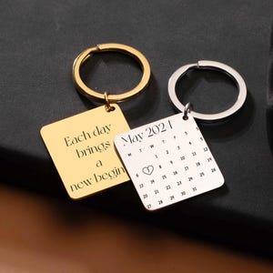 May include: Two square keychains, one gold and one silver, with key rings. The gold keychain reads "Each day brings a new begin." The silver keychain displays a calendar for May 2021 with a heart symbol.