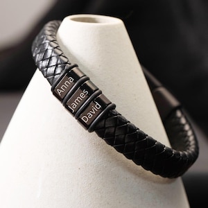 Puede incluir: Pulsera de cuero trenzado negro con tres barras metálicas con los nombres "Anna", "James" y "David". La pulsera se muestra sobre un soporte blanco en forma de cono.