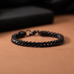 Puede incluir: Una pulsera negra de acero inoxidable con un diseño de cadena trenzada. La pulsera se muestra sobre una superficie marrón con un fondo oscuro. La pulsera mide aproximadamente 20 cm de largo.