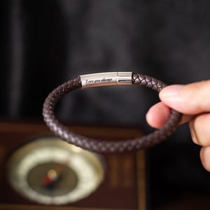 May include: A dark brown, braided leather bracelet with a silver clasp. The bracelet is inscribed with the words "Love you always". The bracelet is made of leather and is a rich, dark brown colour.
