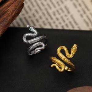 May include: Two snake rings, one silver-toned and one gold-toned, are displayed on a black surface. The silver ring has a textured design with a green gem eye. The gold ring has a similar design. Both rings are coiled in a snake shape.