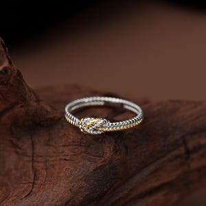 May include: A silver and gold ring with a knot design. The ring has a textured band and a knot at the centre, with silver and gold tones. It is displayed on a dark wooden surface.