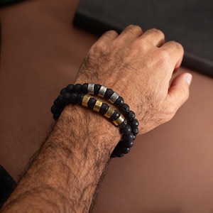 May include: Two black lava stone bead bracelets with personalized name beads in silver and gold. The bracelets are worn on a person's wrist. The background is a solid brown color.