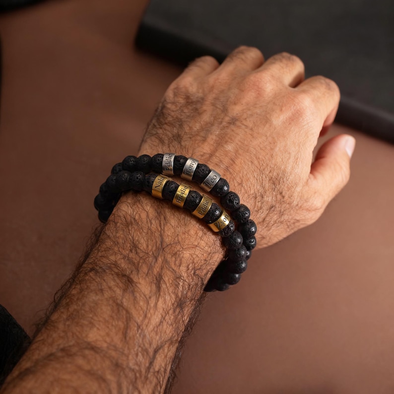 May include: Two black lava stone bead bracelets with personalized name beads in silver and gold. The bracelets are worn on a person's wrist. The background is a solid brown color.