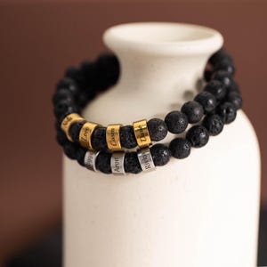 May include: Two black lava stone bracelets with personalized name beads. One bracelet has gold-colored beads with names like "Love" and "Dario." The other bracelet has silver-colored beads with names like "Aron" and "Rivka."