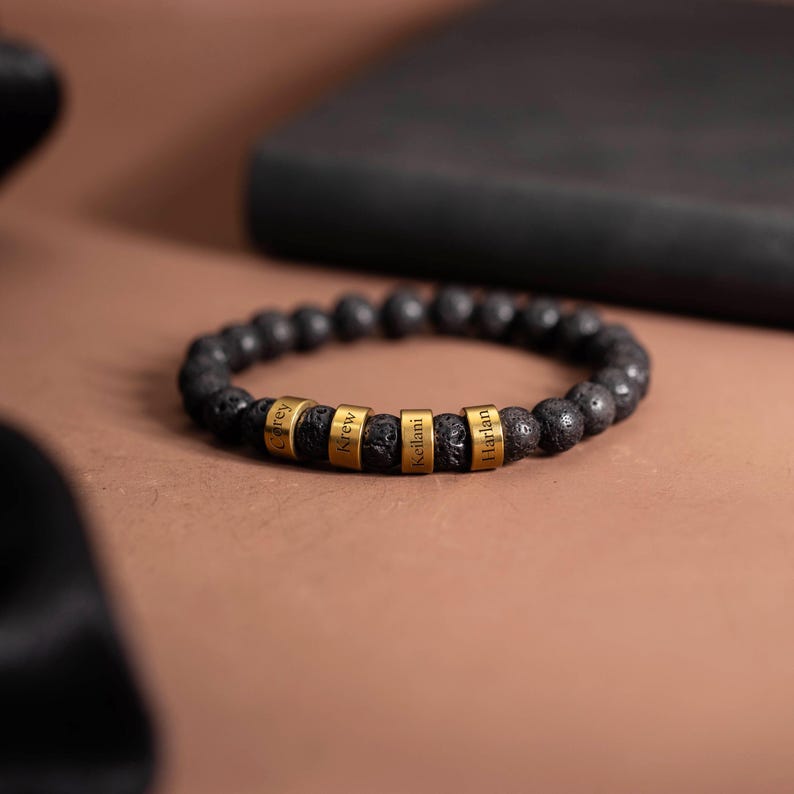 May include: Black lava stone bead bracelet with gold-colored name beads. The bracelet features the names "Carey", "Krew", "Keilani", and "Harlan" engraved on the gold beads. The bracelet is displayed on a brown surface.