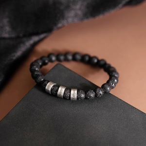 May include: Black lava stone bead bracelet with three silver name beads. The names "Leroy", "Nixon", and "Yahya" are engraved on the silver beads. The bracelet is displayed on a black surface with a brown background.