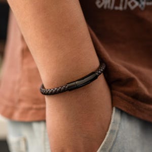 Puede incluir: Una pulsera de cuero trenzado marrón oscuro con un cierre negro. La pulsera se lleva en un brazo, mostrando la textura trenzada y el diseño. El cierre tiene forma rectangular.