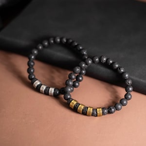 May include: Two black lava stone bead bracelets. One bracelet has silver name rings with the names "Amiri", "Bryson", and "Dilan". The other bracelet has gold name rings with the names "Forest", "Wayne", "Junior", and "Rivka".