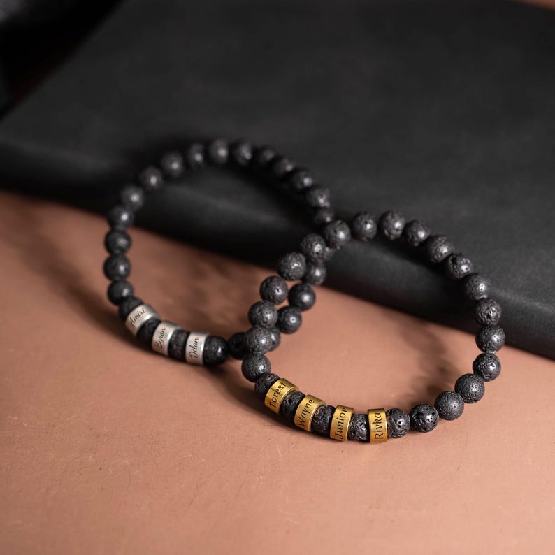 May include: Two black lava stone bead bracelets. One bracelet has silver name rings with the names "Amiri", "Bryson", and "Dilan". The other bracelet has gold name rings with the names "Forest", "Wayne", "Junior", and "Rivka".