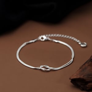 Puede incluir: Una delicada pulsera de plata con un diseño de nudo. La pulsera tiene una fina cadena y una longitud ajustable. El nudo es el punto focal, añadiendo un toque de elegancia. La pulsera se muestra sobre una superficie marrón.
