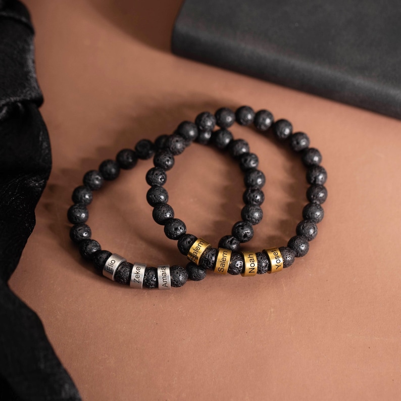 May include: Two black lava stone bead bracelets. Each bracelet has personalized metal beads with names engraved on them. One bracelet has silver beads, the other has gold beads. The bracelets are displayed on a brown surface.