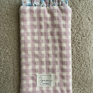 Può includere: Una custodia per telefono rettangolare fatta a mano con un motivo a quadretti rosa e bianco. Il bordo superiore presenta una stampa floreale in blu, rosa e bianco. Una piccola etichetta recita "Izumi Handmade".