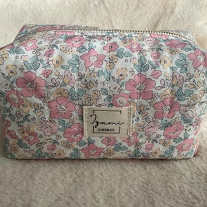 Könnte beinhalten: Eine gesteppte Kosmetiktasche mit einem Blumenmuster in Rosa, Gelb und Blau auf weißem Hintergrund. Die Tasche hat einen Reißverschluss und ein kleines rechteckiges Etikett mit der Aufschrift "Izumi Handmade".
