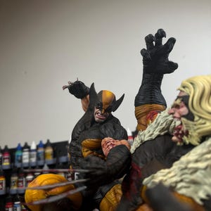 May include: A close-up of two action figures, Wolverine and Sabretooth, in a fighting pose. Wolverine is wearing a yellow and black costume with claws extended, while Sabretooth is wearing a brown and black costume with claws extended. The figures are posed against a backdrop of paint bottles.