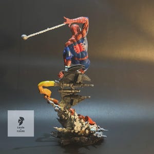 Op de afbeelding: Een rood en blauw Spider-Man-actiefiguur is geposeerd met een web shooter in de hand, gepercht op een zwarte en grijze mechanische basis met een geel en oranje vlam effect. De figuur maakt deel uit van een Lords of Figure-collectie.