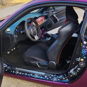 May include: A purple car with black interior and a custom steering wheel. The car has a black and white floral design on the door sill.