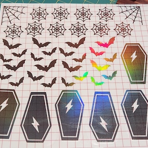 Könnte beinhalten: Halloween-Aufkleberbogen mit Spinnennetzen, Fledermäusen und Särgen. Die Fledermäuse sind braun, rosa und schillernd. Die Särge haben einen Blitz und ein holografisches Finish. Die Aufkleber befinden sich auf einem karierten Hintergrund.