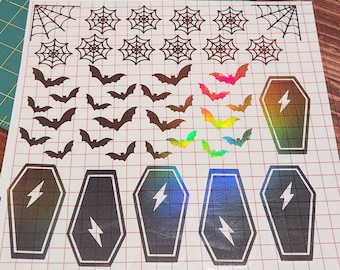 May include: Sheet of Halloween-themed stickers featuring spiderwebs, bats, and coffins. The bats are in various colors, including brown, pink, and iridescent. The coffins have a lightning bolt design and a holographic finish. The stickers are on a grid background.