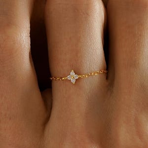 May include: A delicate gold ring with a small, four-pointed starburst design set with tiny gemstones. The ring is on a fine chain. A stylish piece of jewellery. The ring is a beautiful piece of jewelry.