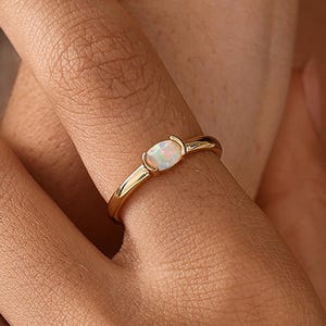 Puede incluir: Un anillo de oro con una pequeña piedra preciosa de ópalo de forma ovalada horizontal, montada en un engaste de bisel liso. La banda es fina, pulida y metálica, mostrando el blanco iridiscente y los sutiles destellos del ópalo central.
