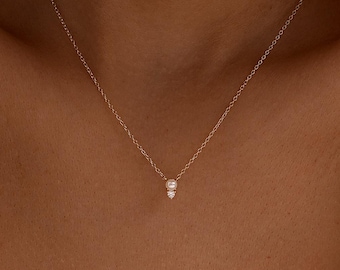 May include: A fine gold necklace with a small pendant featuring two white gemstones. The necklace is worn against a white knitted top. The chain is thin and the pendant is centred.