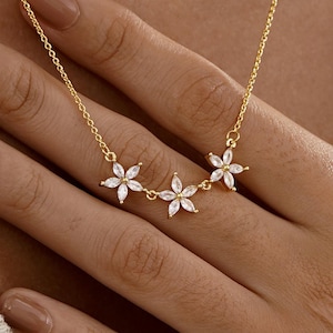 Puede incluir: Un delicado collar dorado con tres colgantes en forma de flor, cada uno adornado con brillantes piedras blancas. El collar se muestra sobre un fondo neutro, resaltando su elegante diseño. La cadena es fina y las flores son simétricas.