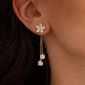 Pode incluir: Brincos de flores douradas com pedras preciosas quadradas penduradas. Os brincos apresentam um design de tachinha floral com uma corrente que suspende duas pedras preciosas quadradas. Os brincos est&atilde;o em um fundo neutro.