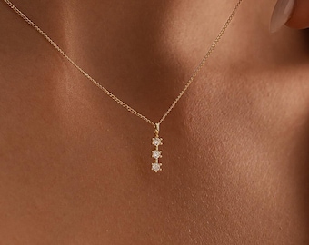 May include: A delicate gold necklace featuring a pendant with three small, sparkling gemstones. The necklace has a fine chain and is worn around the neck. The gemstones are set in a vertical line, adding a touch of elegance to the jewelry.