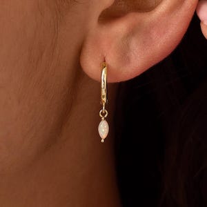 Könnte beinhalten: Ein goldener Creolen-Ohrring mit einem kleinen, baumelnden, marquiseförmigen Opal-Anhänger. Der Ohrring ist am Ohrläppchen befestigt und zeigt einen polierten Goldreif sowie einen glatten, schillernden weißen Opalstein, der das Licht einfängt.
