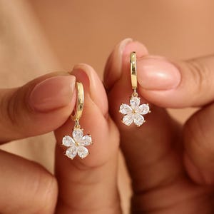 May include: A pair of gold hoop earrings, each featuring a dangling flower-shaped charm. The flower charms are composed of five pear-cut clear white stones arranged in a petal pattern, with a central stone accentuating the floral design.
