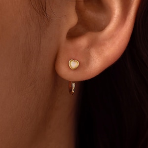 Può includere: Un orecchino a cerchio dorato con un piccolo ciondolo a forma di cuore con un inserto bianco iridescente. L'orecchino è fissato al lobo dell'orecchio, mostrando una finitura metallica lucida e un design delicato che cattura la luce.