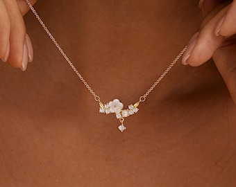 May include: A delicate gold necklace featuring a floral design with white petals and sparkling accents. The necklace has a fine chain and is adorned with small, clear gemstones. The floral pendant is centered on the chain.