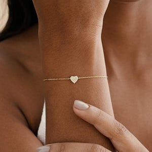 May include: A delicate gold bracelet featuring a heart-shaped charm. The charm is encrusted with small, sparkling stones. The bracelet is worn on a wrist, showcasing its fine chain and the heart pendant.
