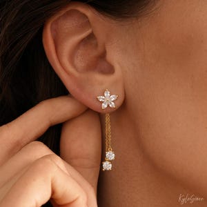 Pode incluir: Brincos de bot&atilde;o dourados em forma de flor com pedras de zirc&ocirc;nia c&uacute;bica penduradas. Os brincos apresentam um design floral no l&oacute;bulo da orelha, com uma corrente que suspende duas pedras redondas e transparentes.