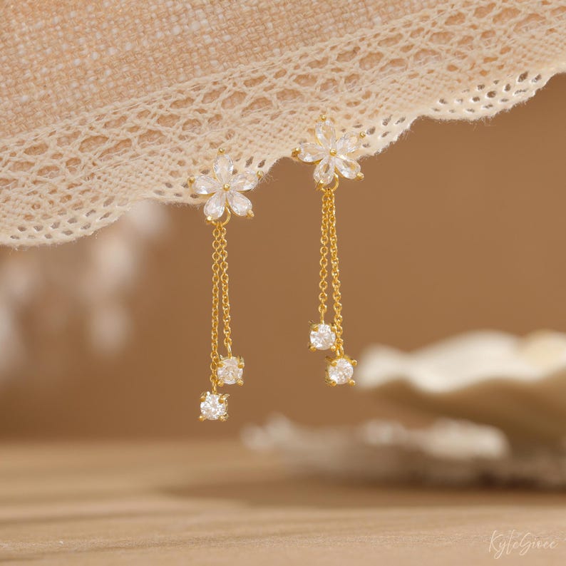 Pode incluir: Um par de brincos dourados com um design floral no topo e correntes penduradas. Cada corrente tem duas pedras preciosas redondas e transparentes. Os brincos s&atilde;o exibidos em um fundo macio e neutro.