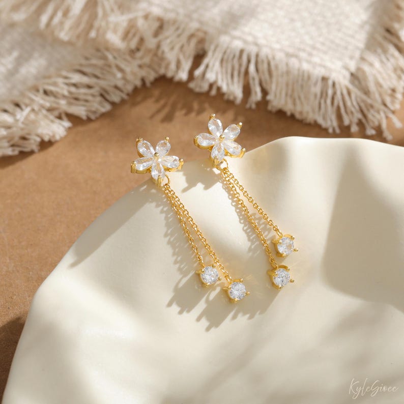 Pode incluir: Brincos pendurados dourados com design floral e pedras transparentes. Correntes delicadas pendem, cada uma com uma pedra redonda e transparente. Os brincos s&atilde;o exibidos em uma superf&iacute;cie branca e curva.