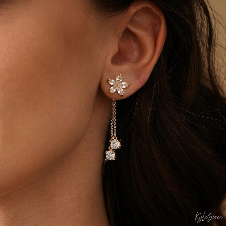 Pode incluir: Brincos de flores douradas com cristais transparentes pendurados. Os brincos apresentam um pino em forma de flor com uma corrente que suspende dois cristais redondos e brilhantes. O design &eacute; elegante.
