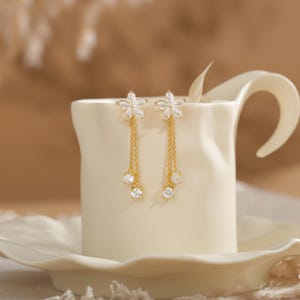 Pode incluir: Um par de brincos dourados com um design floral. Cada brinco apresenta um topo em forma de flor com pedras claras e brilhantes e correntes penduradas com tr&ecirc;s pedras redondas e claras. Os brincos s&atilde;o exibidos em uma superf&iacute;cie de cer&acirc;mica branca.