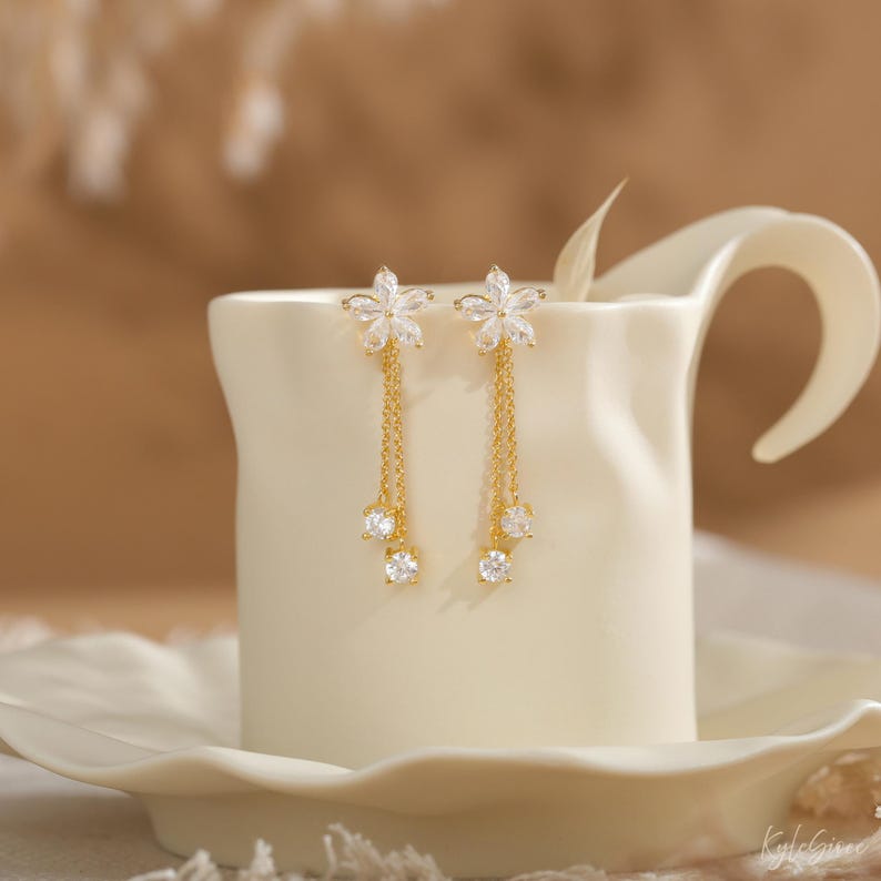 Pode incluir: Um par de brincos dourados com um design floral. Cada brinco apresenta um topo em forma de flor com pedras claras e brilhantes e correntes penduradas com tr&ecirc;s pedras redondas e claras. Os brincos s&atilde;o exibidos em uma superf&iacute;cie de cer&acirc;mica branca.