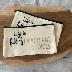 Könnte beinhalten: Zwei Canvas-Reißverschlusstaschen mit schwarzem Besatz und dem Text "Life is full of IMPORTANT CHOICES" in Schwarz und Beige. Die Taschen liegen auf einem Holztablett.
